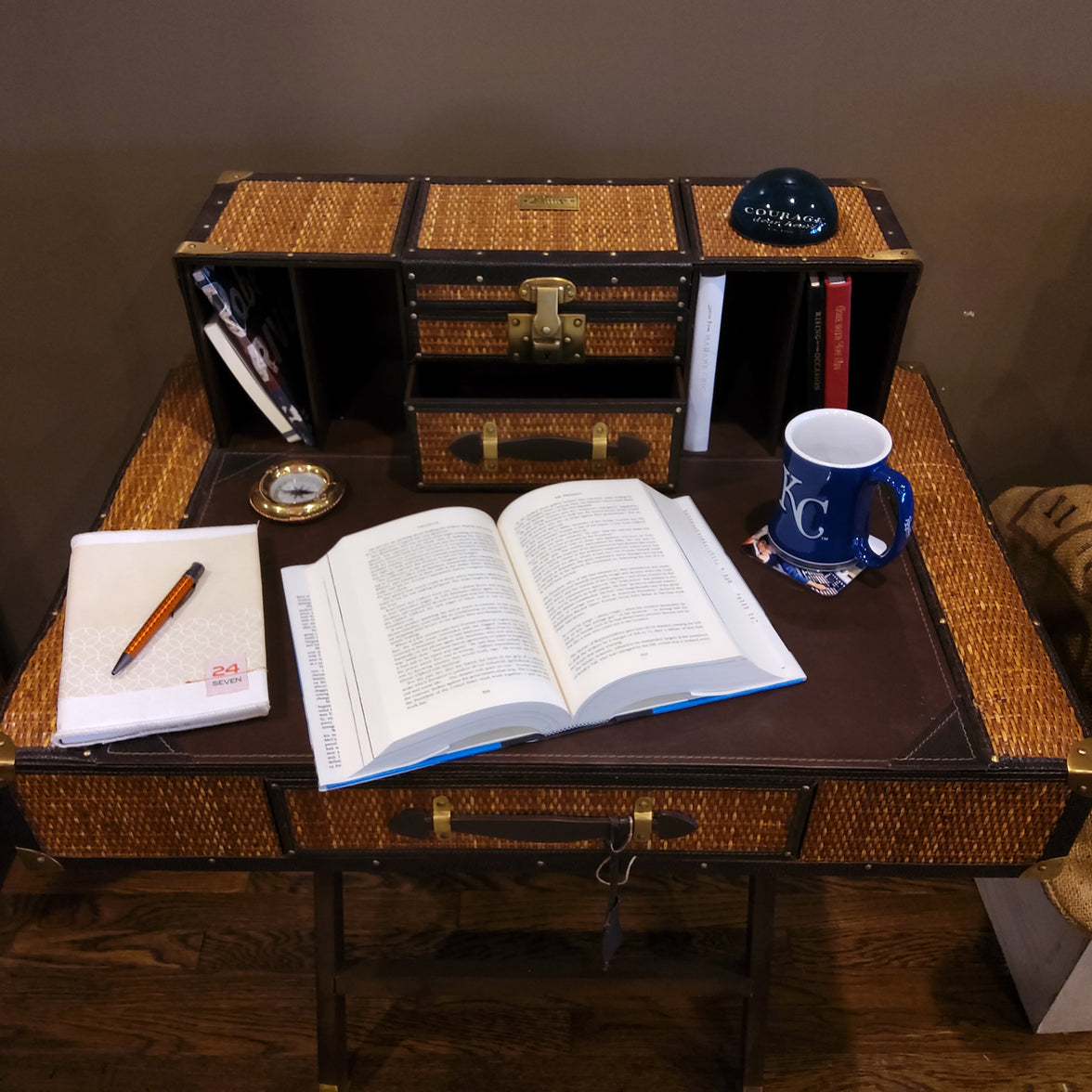 A traditional style campaign desk with a wicker surface, leather-bound books, a coffee mug, and a small lamp, set against a brown backdrop.