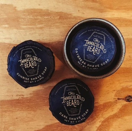 Three Immaculate Beard shave soaps in their black paper wrappings on a wooden background, with one of them in a handmade stoneware shave soap bowl.