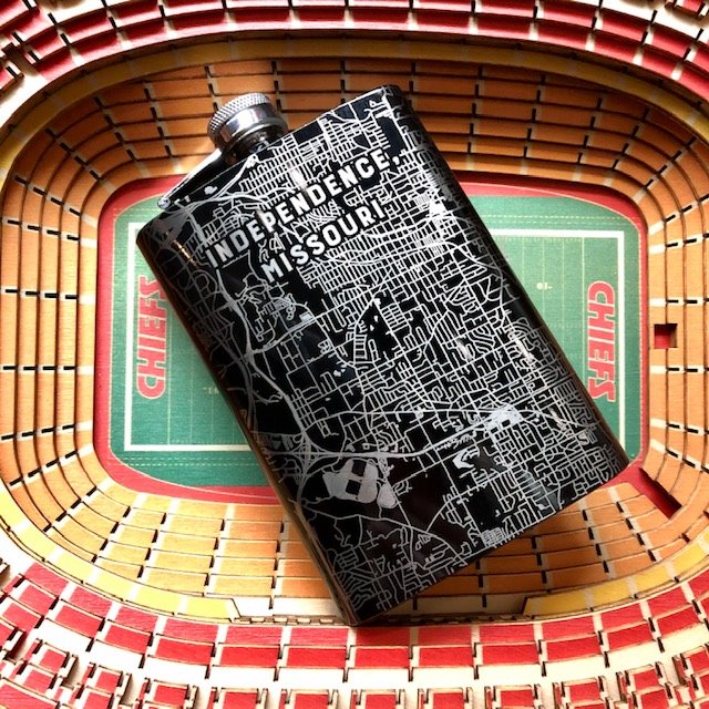 A black and silver flask with an etched map of Independence, Missouri on a wooden replication of Arrowhead Stadium.