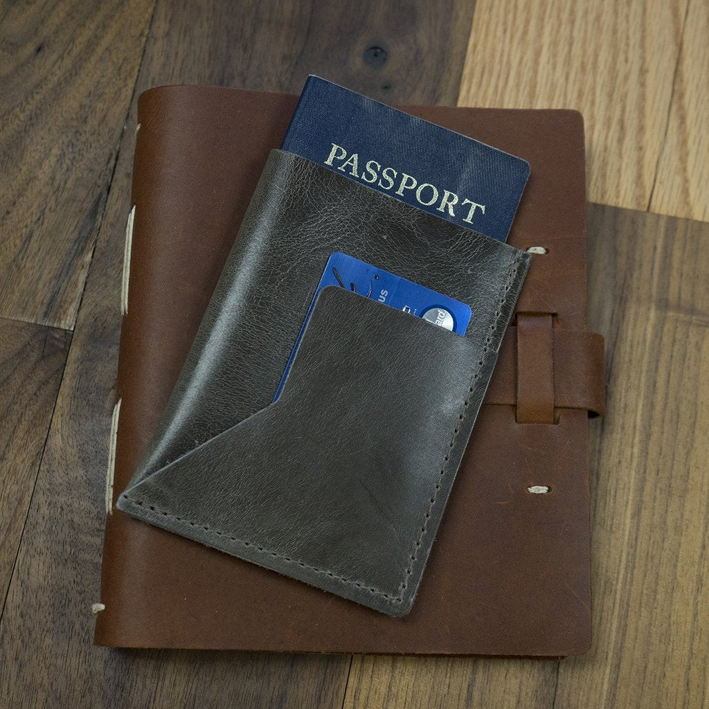 A black leather passport sleeve with a passport and credit card inside, lying on a leather journal on a wooden surface.