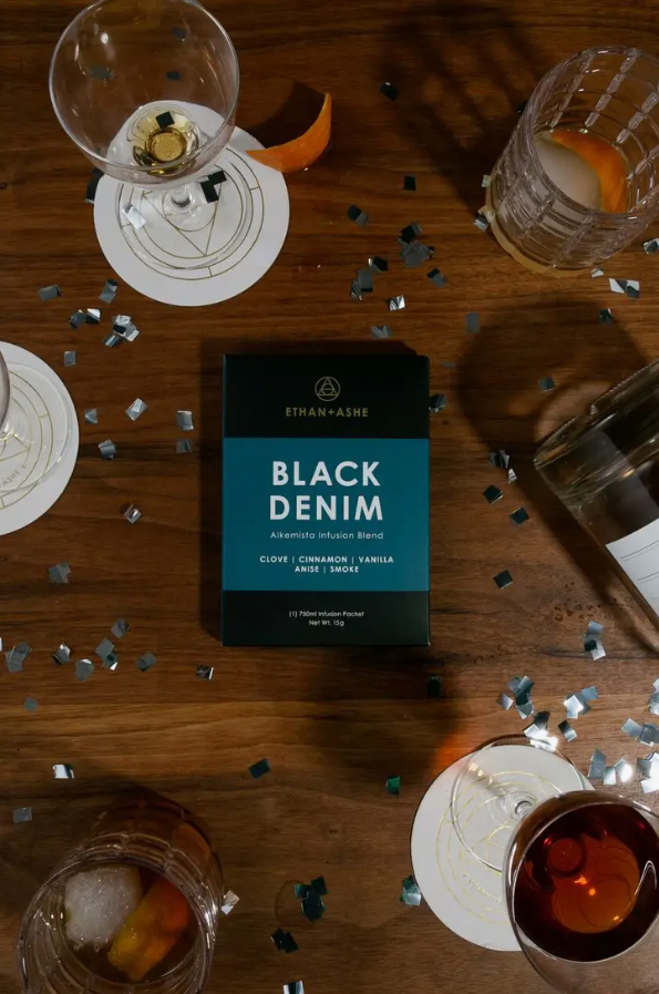 A package of 'Black Denim' Alkemista Infusion Blend by Ethan+Ashe displayed on a wooden table with drinks placed around it.