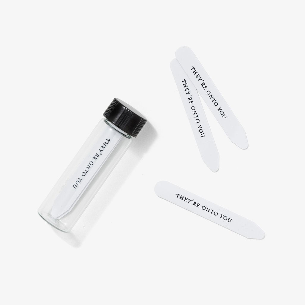 A set of clear collar stays in a glass vial with the phrase 'THEY'RE ON TO YOU' printed on each stick.