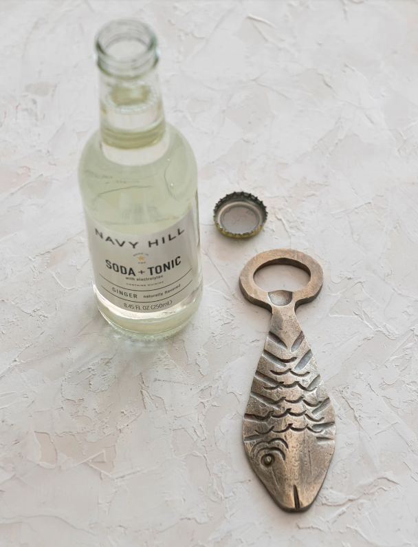A metal fish-shaped bottle opener laying on a textured surface next to a bottle cap and an open body of soda and tonic.
