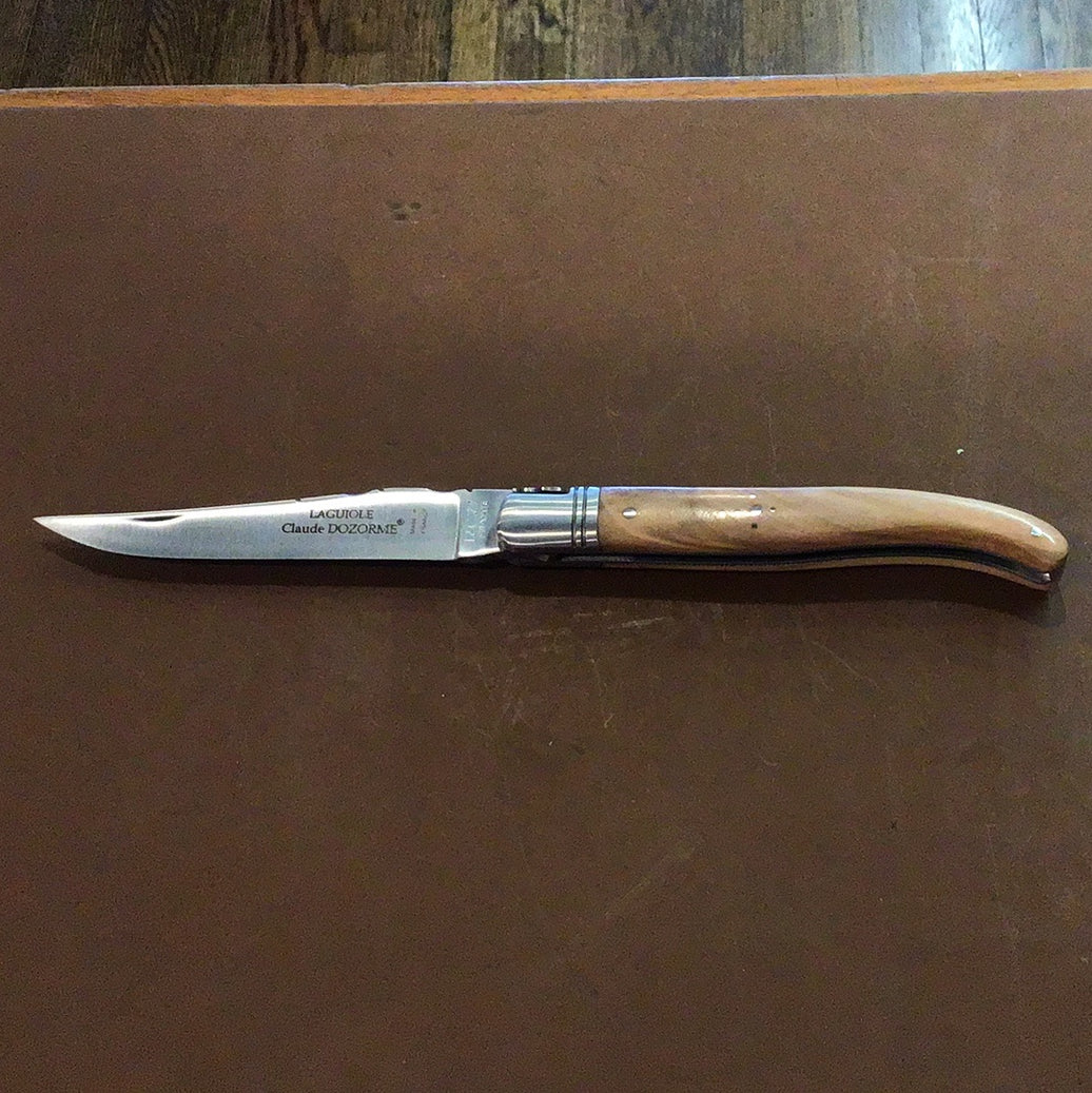 A Claude Dozorme Laguiole pocket knife with a light brown wooden handle and a stainless steel blade, displaying a mythical bee design near the handle's sheath.