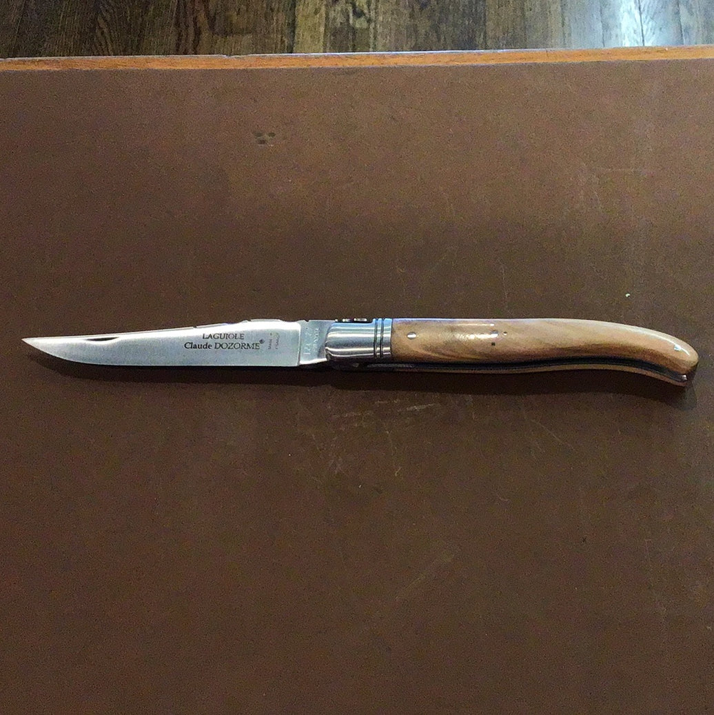 A Claude Dozorme Laguiole pocket knife with a light brown wooden handle and a stainless steel blade, displaying a mythical bee design near the handle's sheath.
