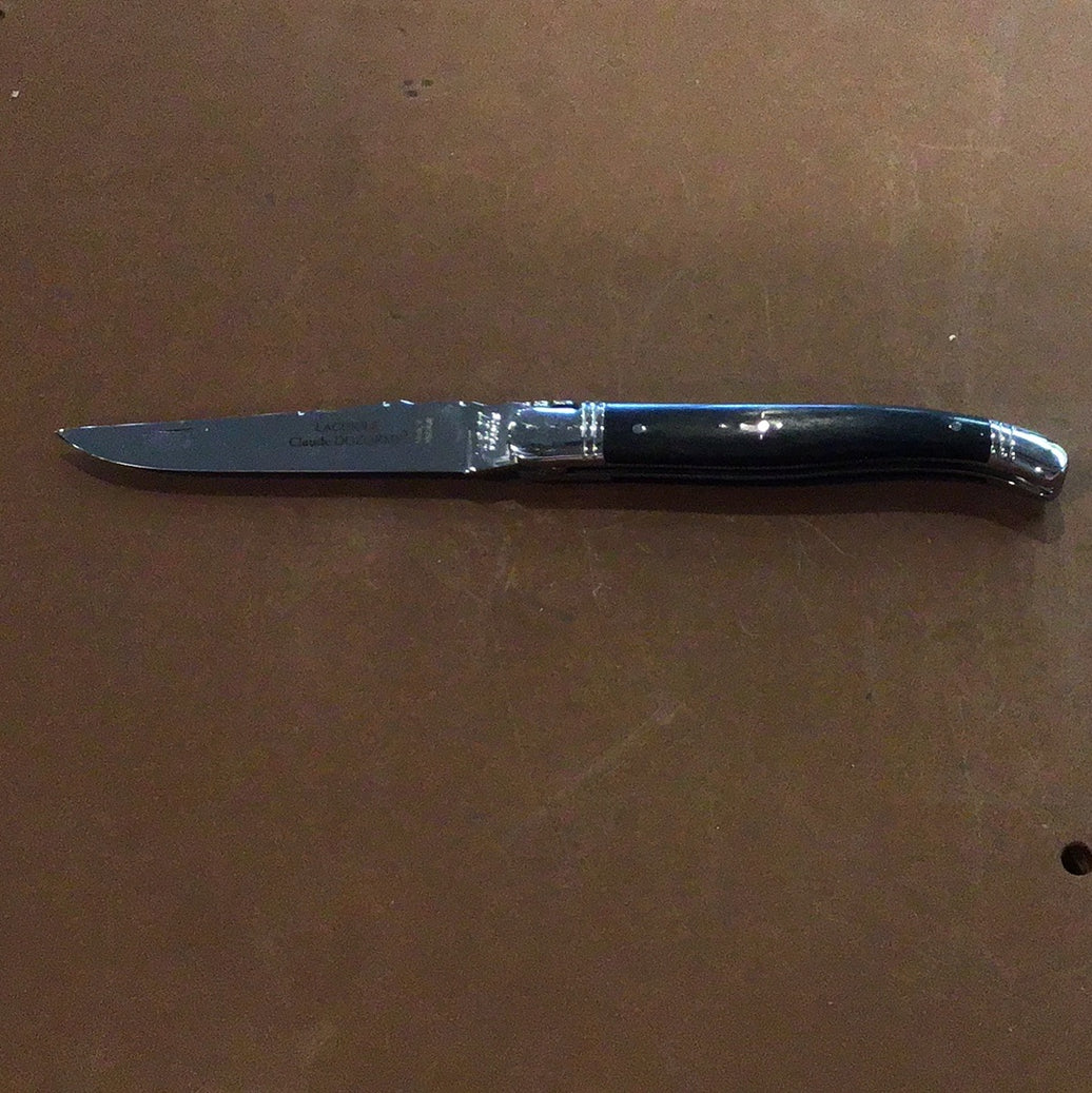 A Claude Dozorme Laguiole pocket knife with a black handle and a curved Damascus steel blade, displayed on a textured background.