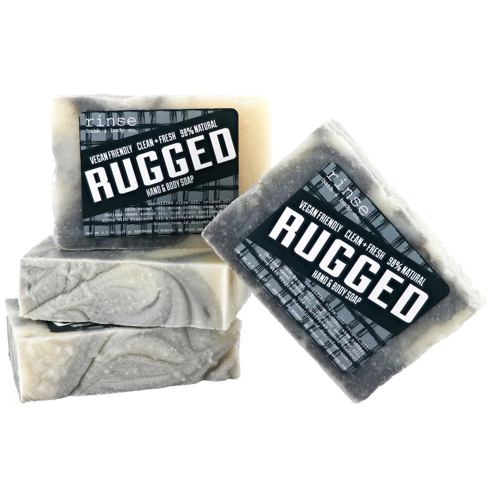 A packaged bar soap labeled 'RUGGED' by Rinse bath & Body, with a grayish color and activated charcoal visible on the surface.