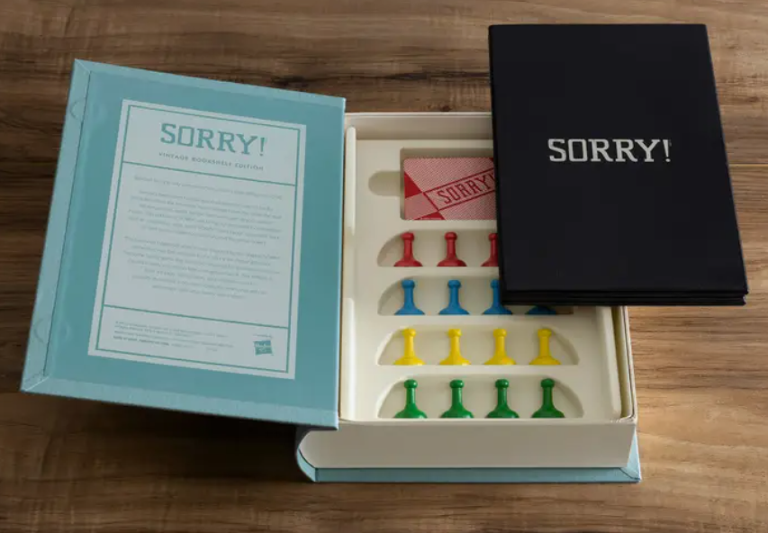 A vintage, book-shaped package edition of the board game SORRY! open to show the pieces and the folded up game board on a wooden surface. 
