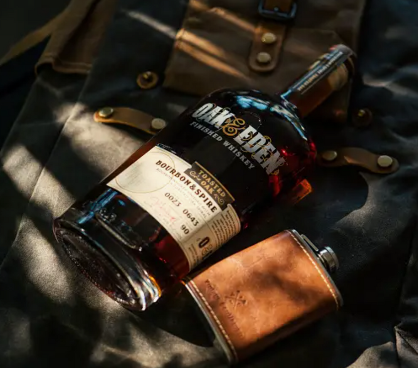A Vintage Gentlemen leather flask  lying on a backpack next to a bottle of Oak & Eden finished whiskey.