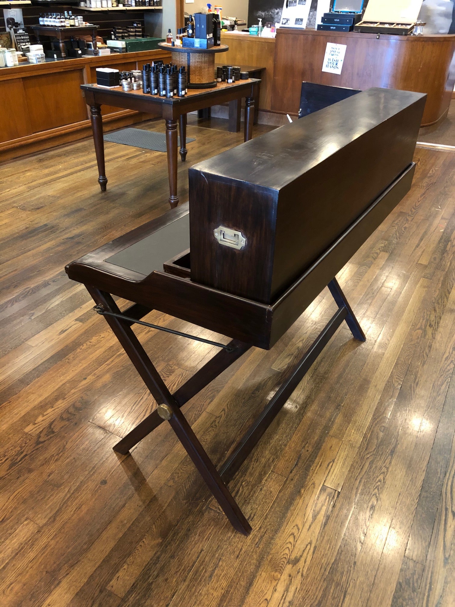 Side/back view of a dark wood writing desk with folding legs and a removable mail slot/shelf unit. It is displayed in a room with various books and office supplies.