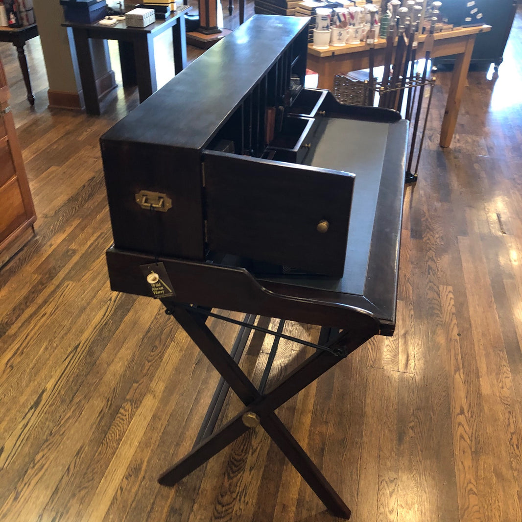 Side view of a dark wood writing desk with folding legs and a removable mail slot/shelf unit. It is displayed in a room with various books and office supplies.